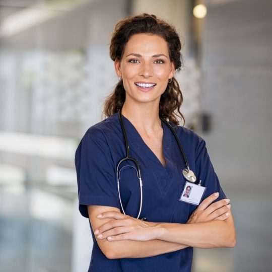 Portrait of happy young female nurse with folded arms standing in hospital hallway. Confident doctor woman in uniform and stethoscope looking at camera with copy space. Portrait of beautiful young healthcare worker working in private clinic.